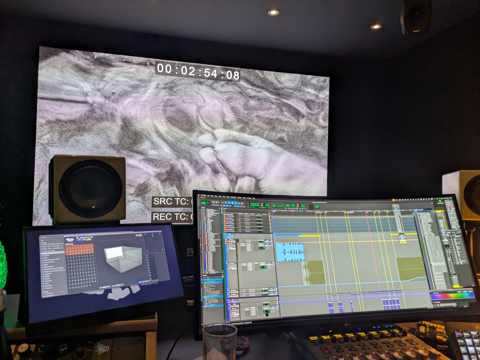 A dubbing studio in low light. A grainy black-and-white film frame is projected on the far wall with burnt-in timecode reading 00:02:54:08. In front of it, a Pro Tools session fills a wide curved monitor, a Dolby Atmos panner sits on a second screen, and a mixing desk and monitor speakers surround the desk.