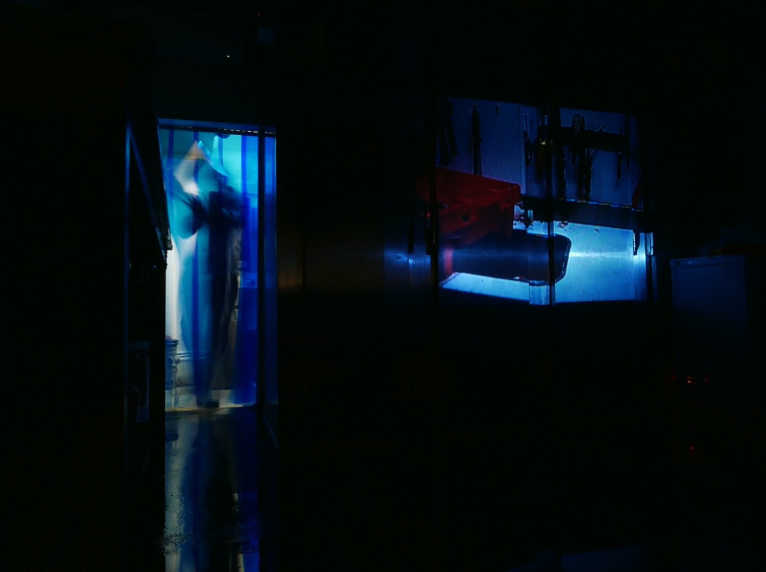 A dark fish market at night. A figure in overalls passes through blue-lit plastic strip curtains in a doorway, reflected in the wet floor; tools and red machinery are visible in a second lit doorway across the room.