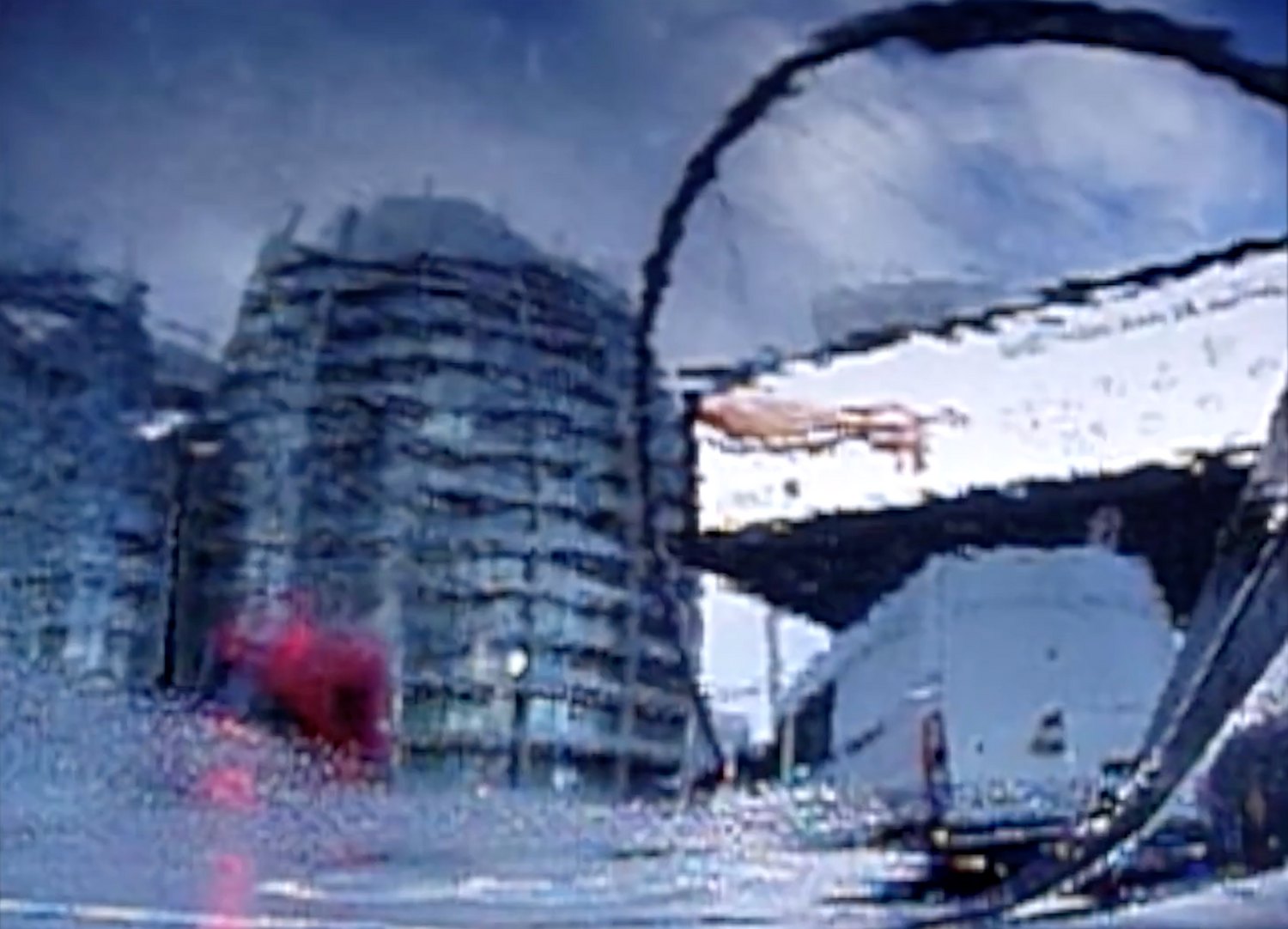 A smeared, degraded video image of a city under construction: scaffolded tower blocks under a grey sky, a dark curved form arcing across the frame, a blurred red mass at street level.